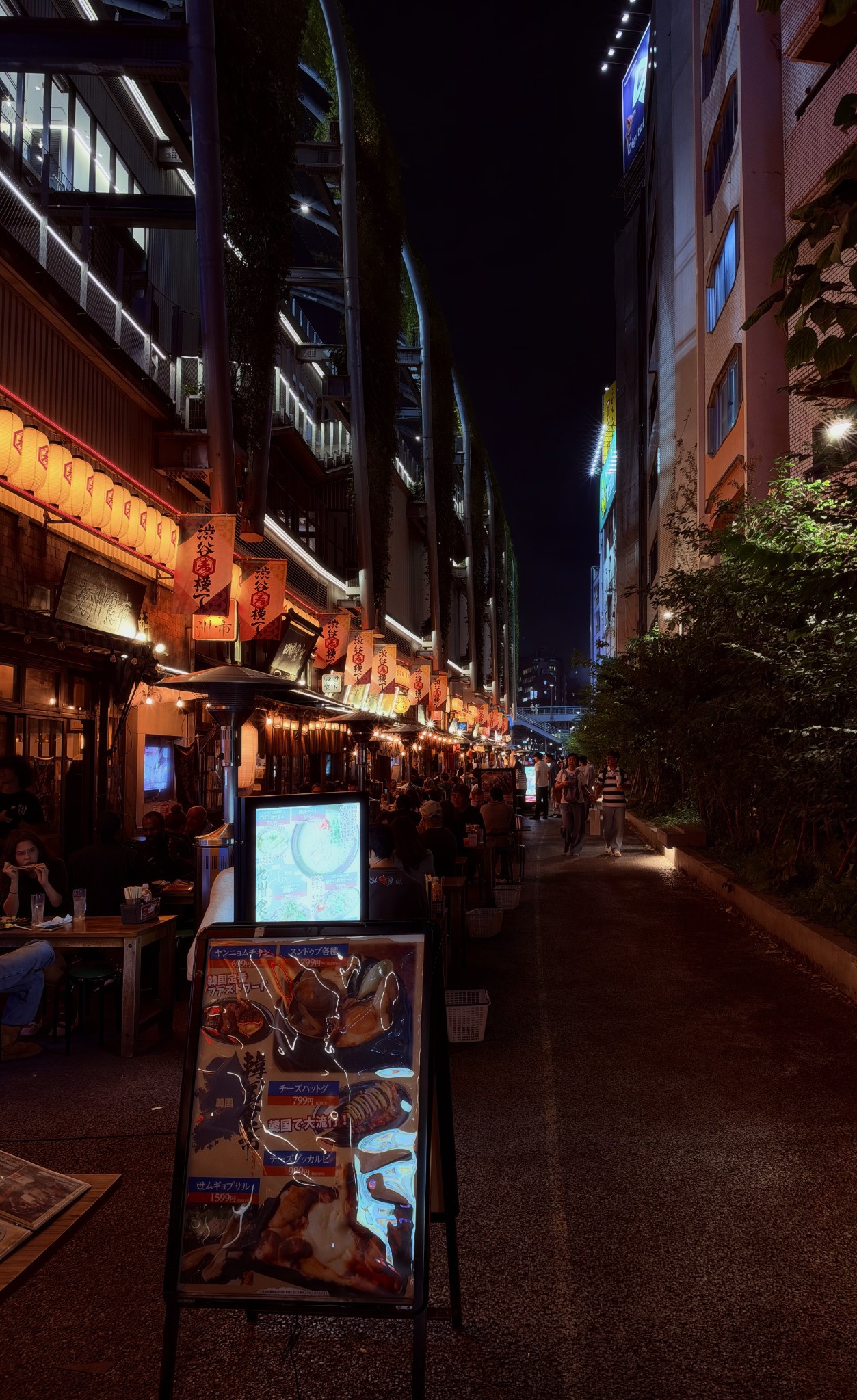 Lantern-lit restaurants and closely spaced outdoor tables at Shibuya Yokocho, beneath tall buildings at night.