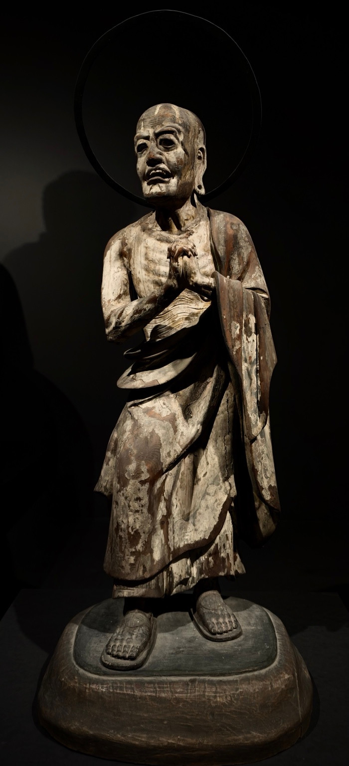 A weathered robed statue with joined palms and a circular halo, lit against a dark background.