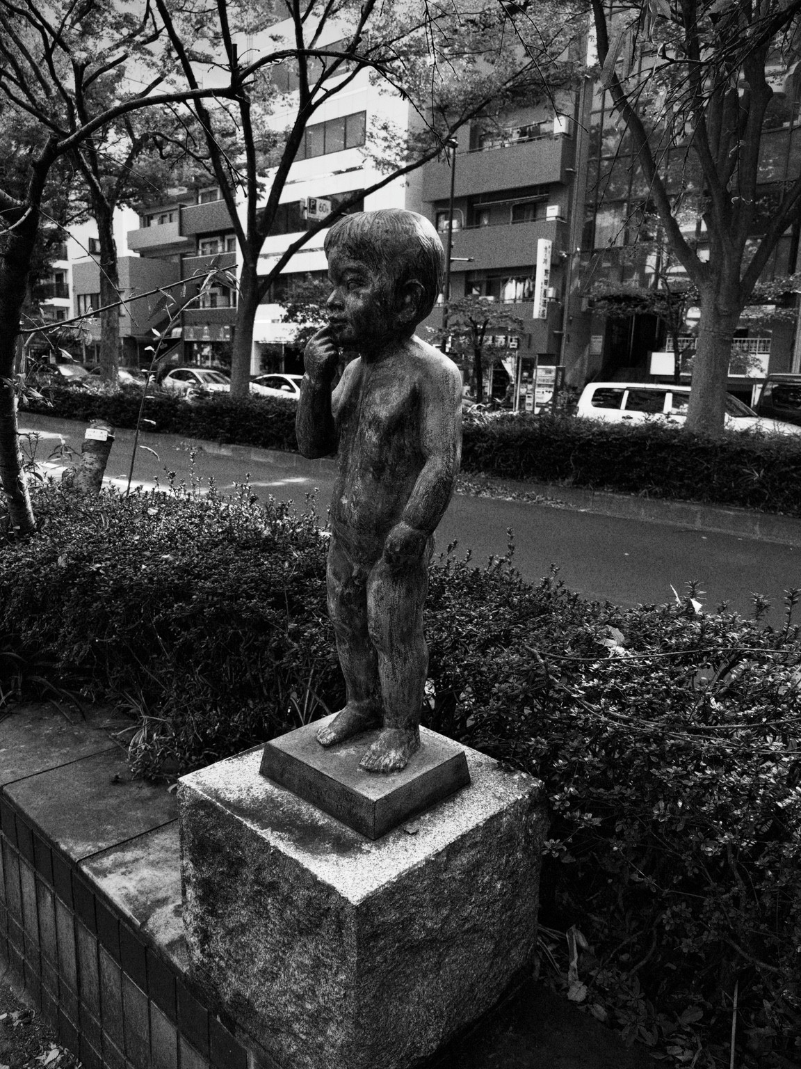 Black-and-white photograph of a child sculpture among shrubs beside a city street.