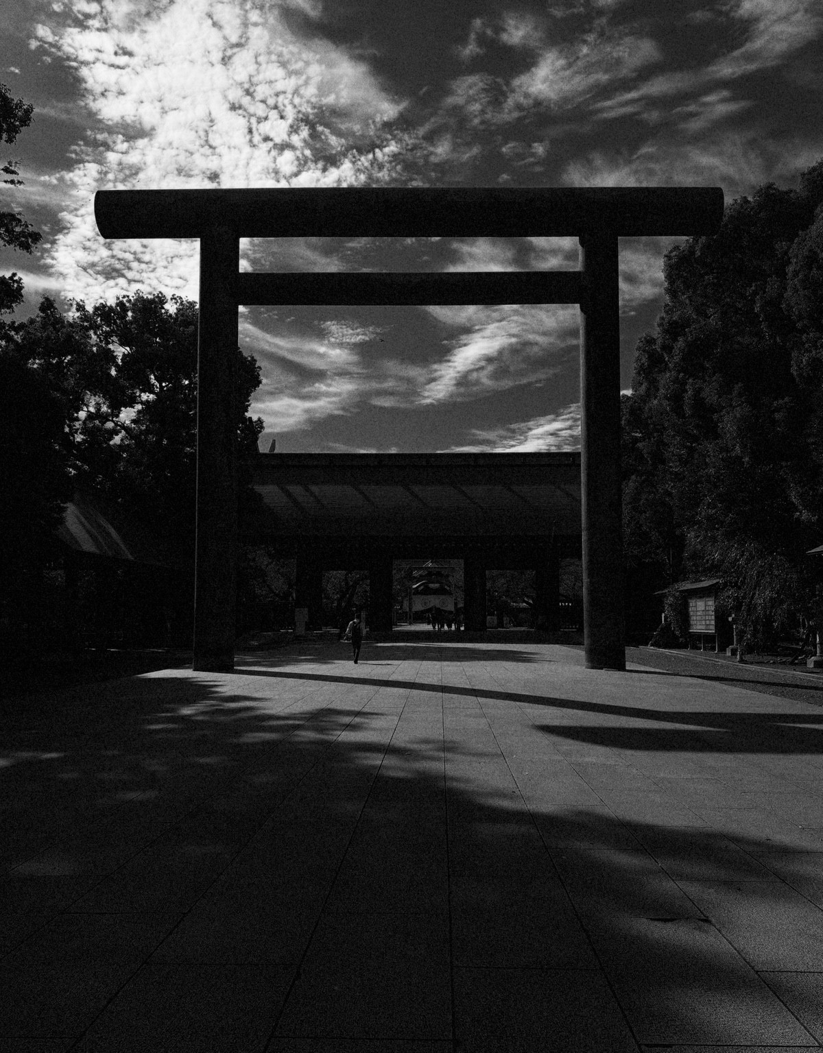 A large torii gate and shrine entrance beneath a cloud-filled sky, photographed in black and white.
